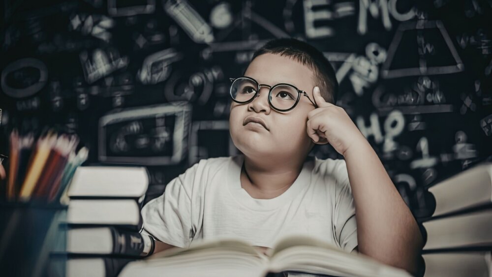 A young boy sitting at a desk in front of a blackboard filled with  mathematical equations. He is thinking and looking up toward the ceiling with his finger on his temple, while his books a spread out in front of him.