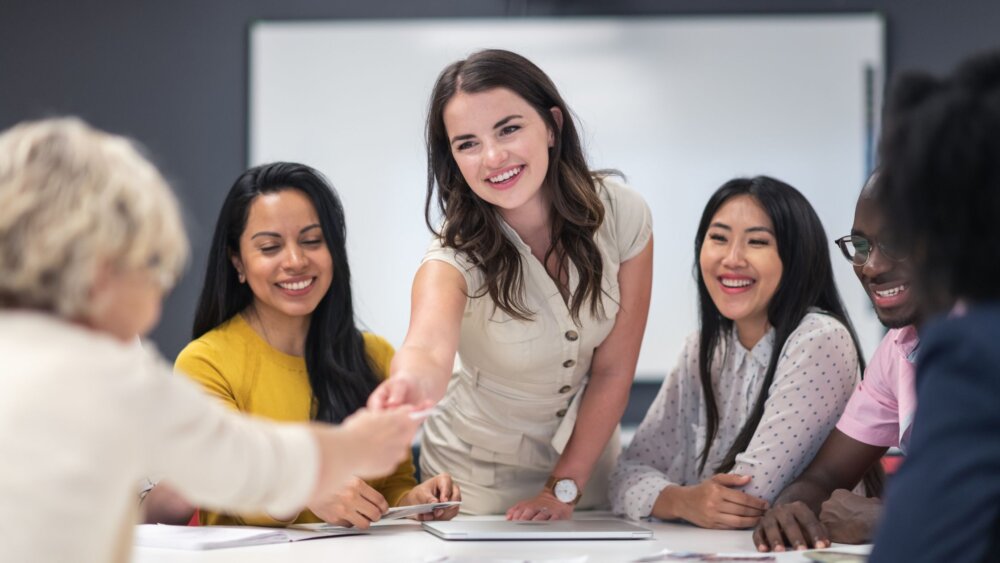 A business woman leans across a desk to shake the hand of new person she has chosen to introduce to her team